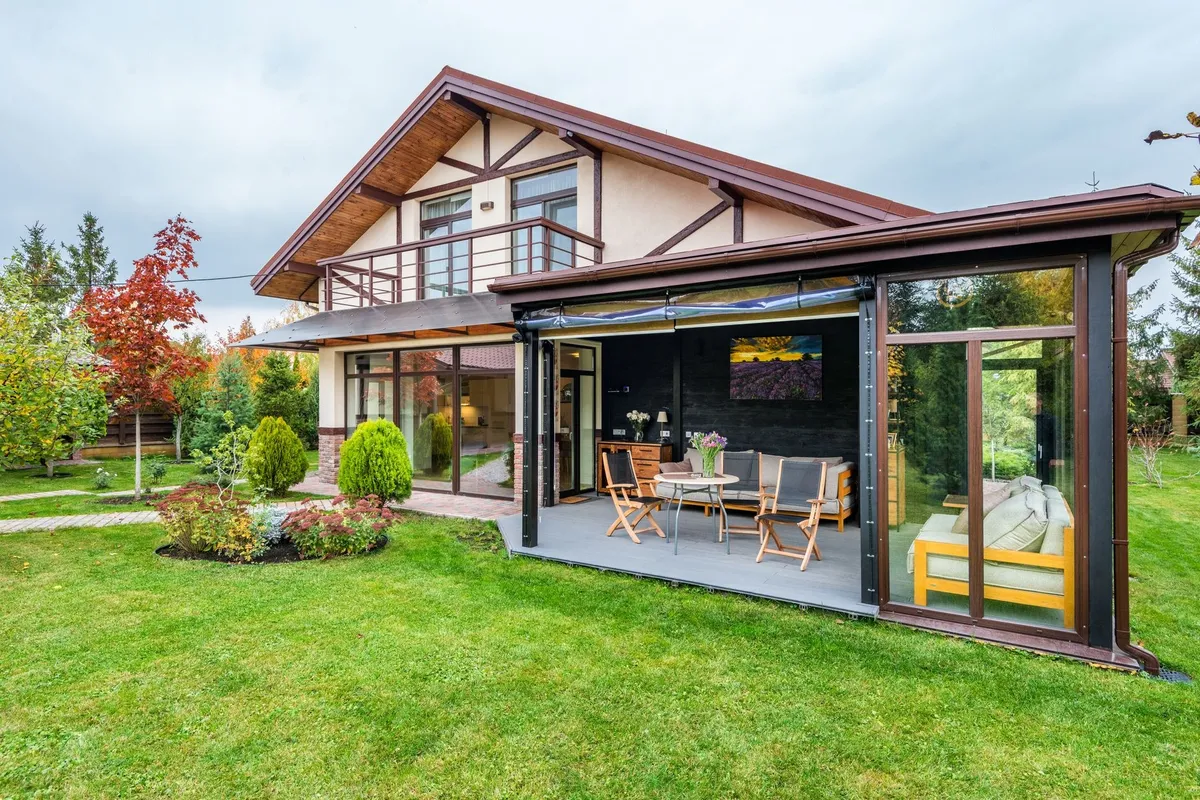 A cozy residential cottage with a balcony and open veranda overlooking a lush lawn and trees.